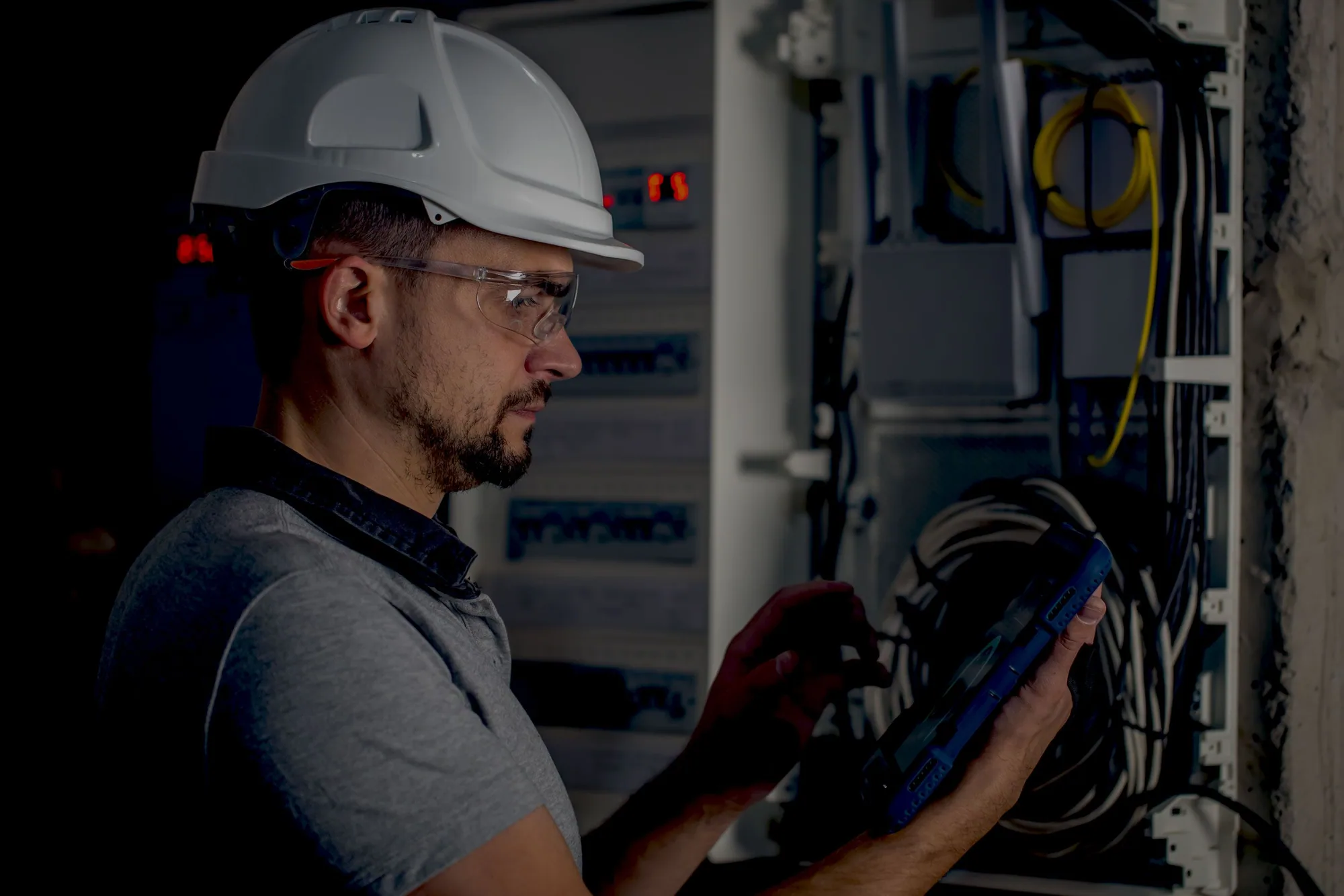 A technician in a hard hat and safety glasses working at an open electrical cabinet in low light, a rugged tablet in one hand.