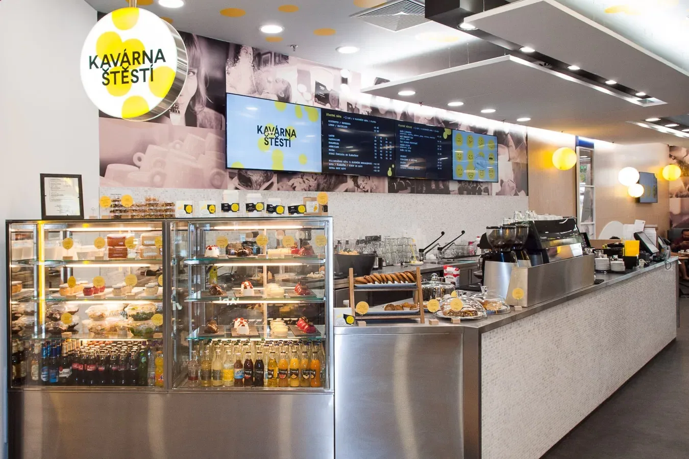 The counter of a Lucky Café: pastry cabinets and a coffee machine under menu screens, with the yellow “Kavárna štěstí” logo hanging above.