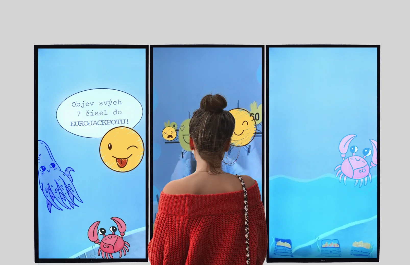 A person standing before three large touchscreen panels illustrated with cartoon sea creatures and Czech Eurojackpot lottery messaging, part of an interactive café installation.