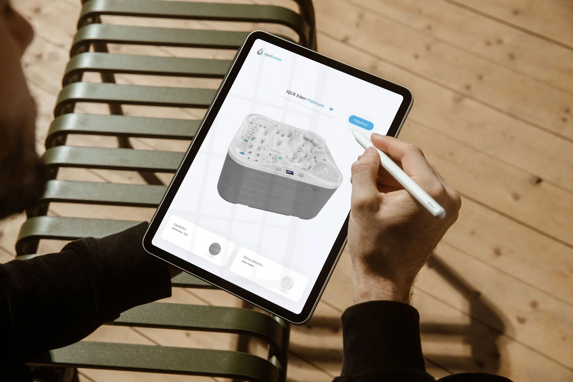 A hand holding an Apple Pencil over an iPad showing the Spahouse 3D configurator: a rendered hot tub model with cladding and surface-color options and an order button, resting on an outdoor deck chair.